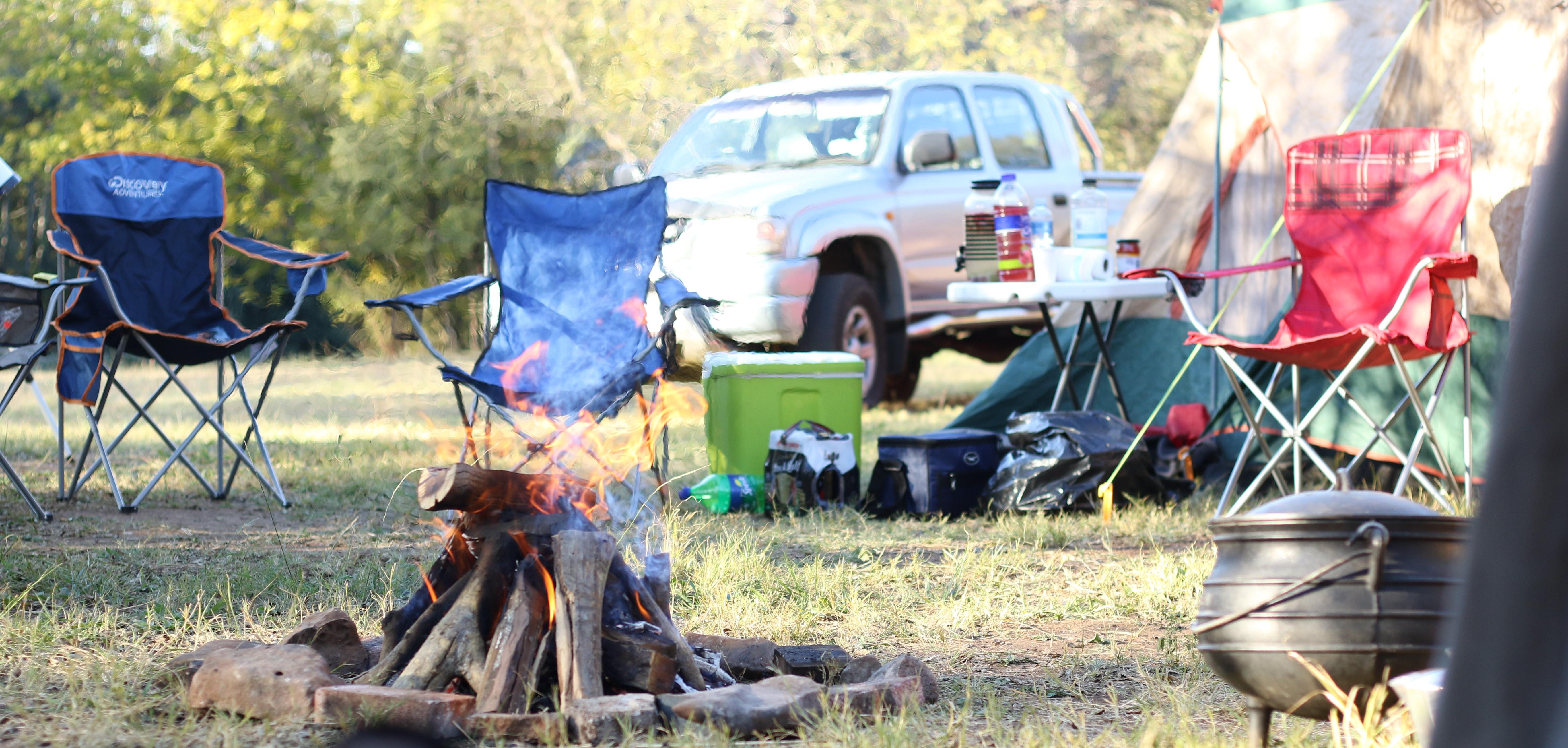 Stock photo of a camp with fire, camping chairs, the side of a tent, and a silver pickup truck in the background