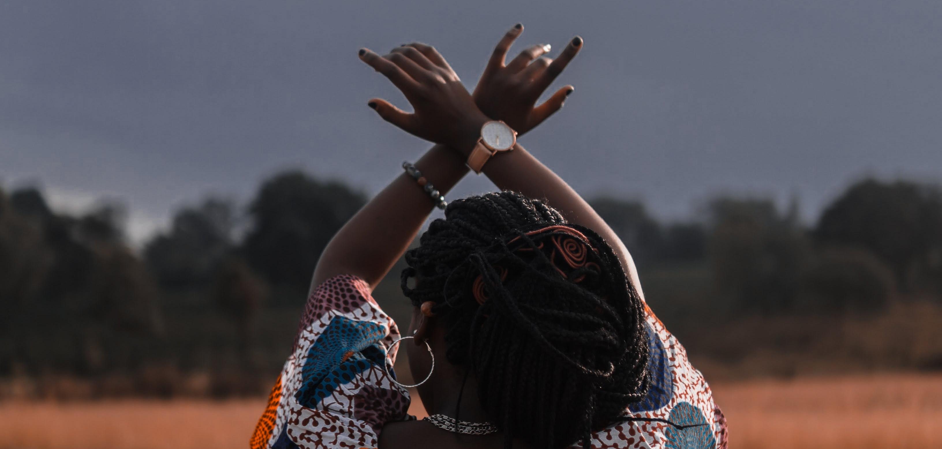 Black woman in field facing away from camera with arms in the air crossed at the wrist. 