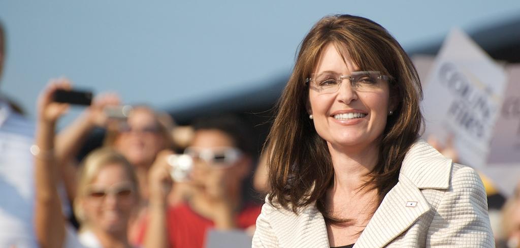 Sarah Palin in a white jacket, in front of a crowd, smiling.