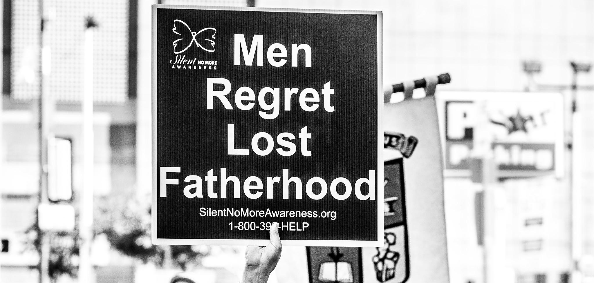 A man wearing sunglasses holding a sign that says "Men Regret Lost Fatherhood"