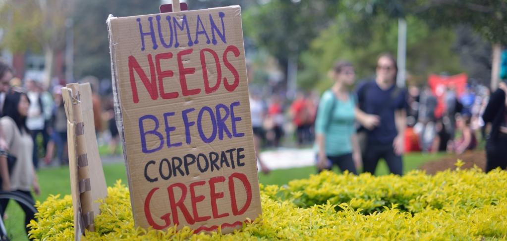A cardboard sign that says Human Needs Before Corporate Greed