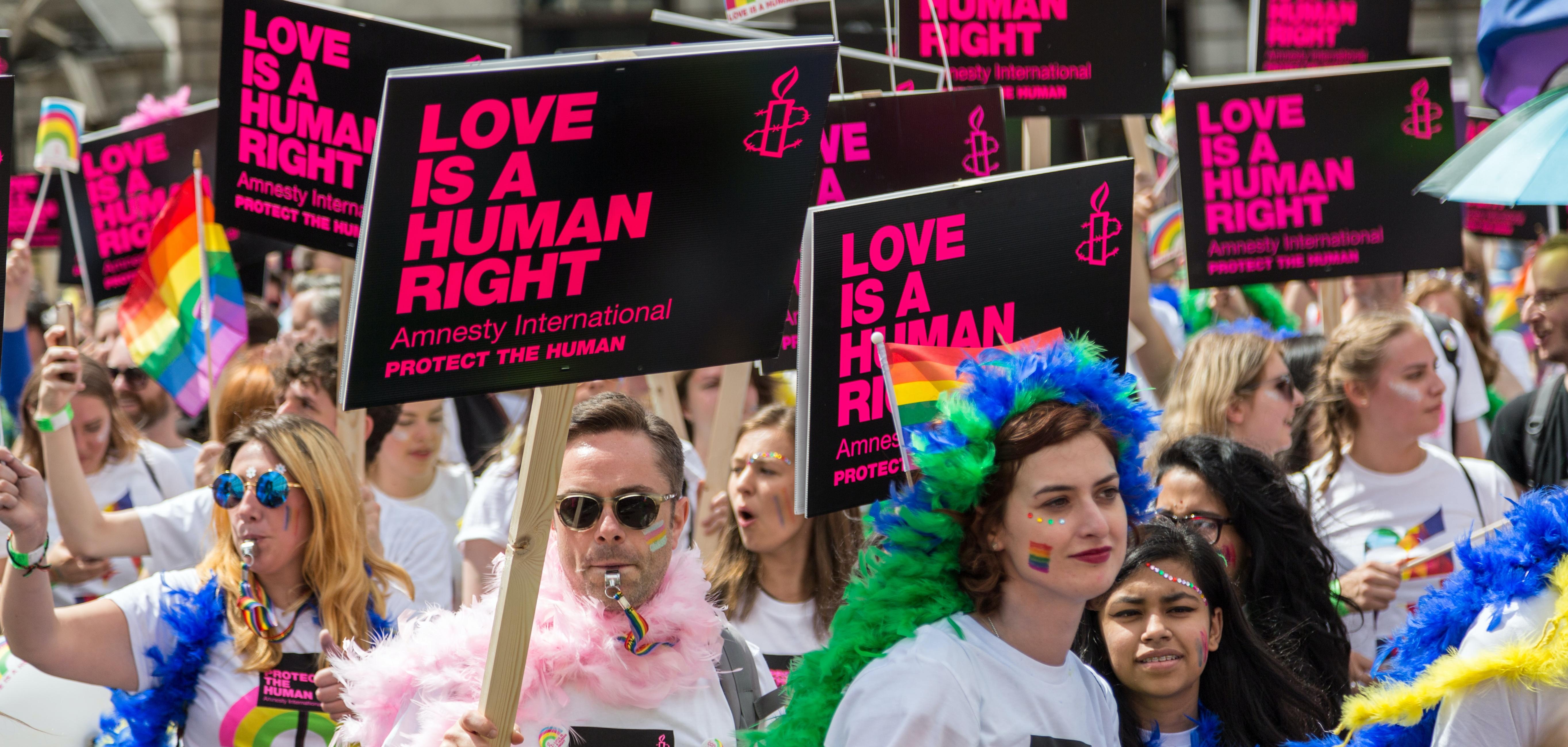 People in a protest wearing rainbow wigs, holding signs that say "love is a human right"