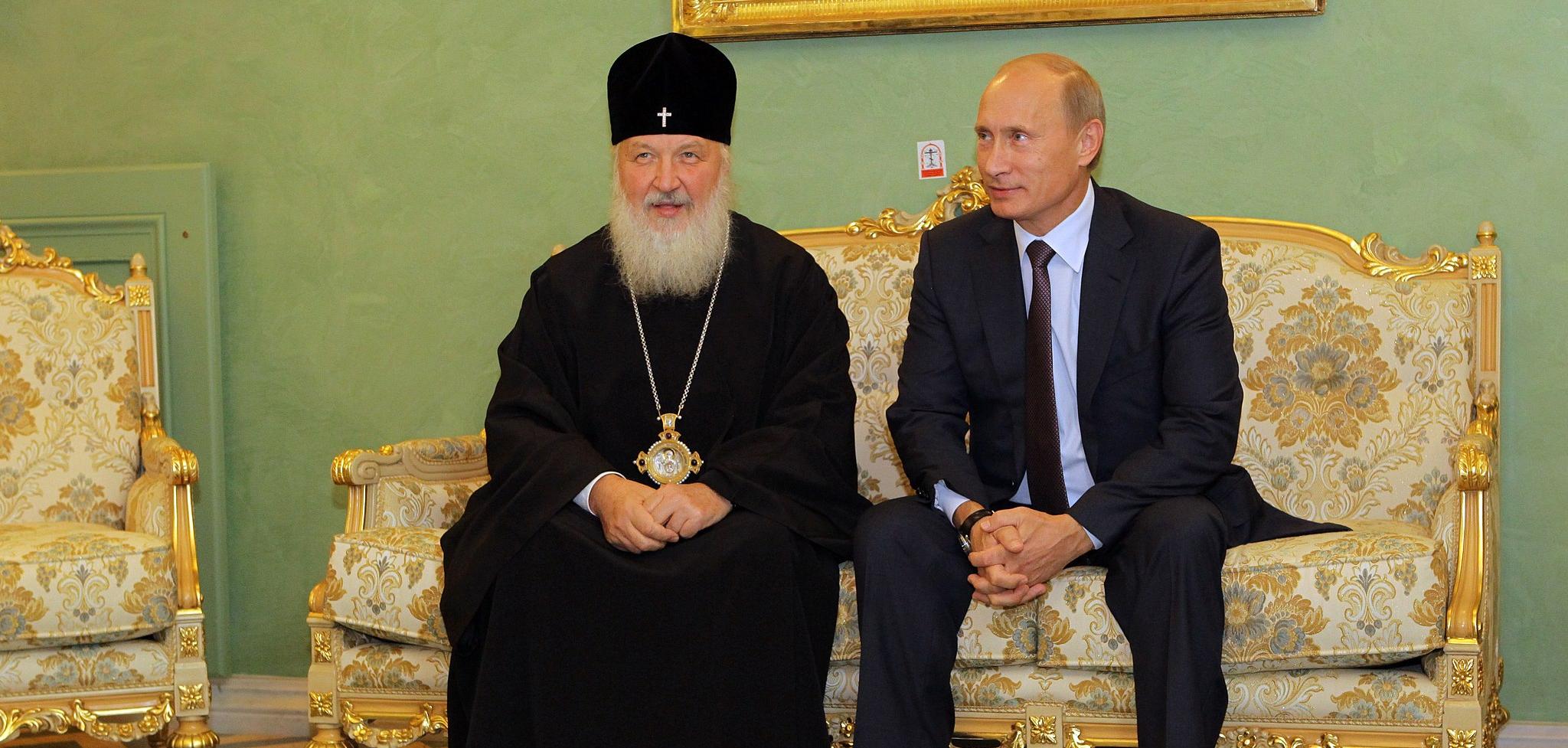 A man in a black robe and a black hate with a cross on it sitting next to Vladimir Putin on a golden couch.