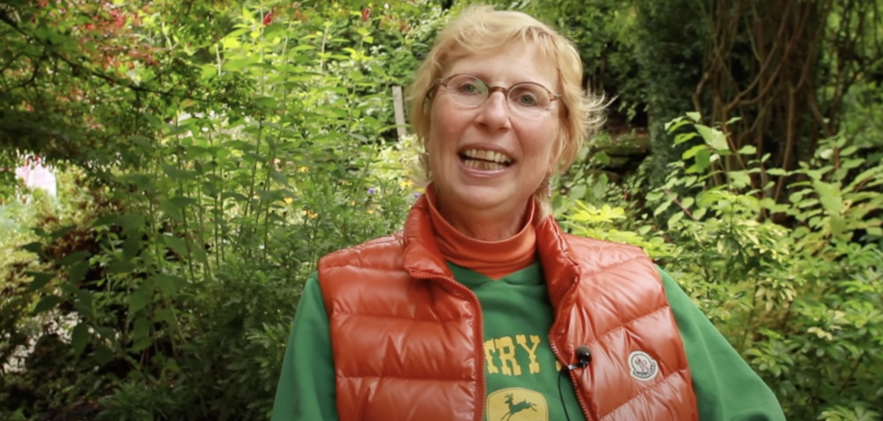 A White woman with short blond hair wearing a green t-shirt and orange sleeveless jacket sitting in front of some bushes.