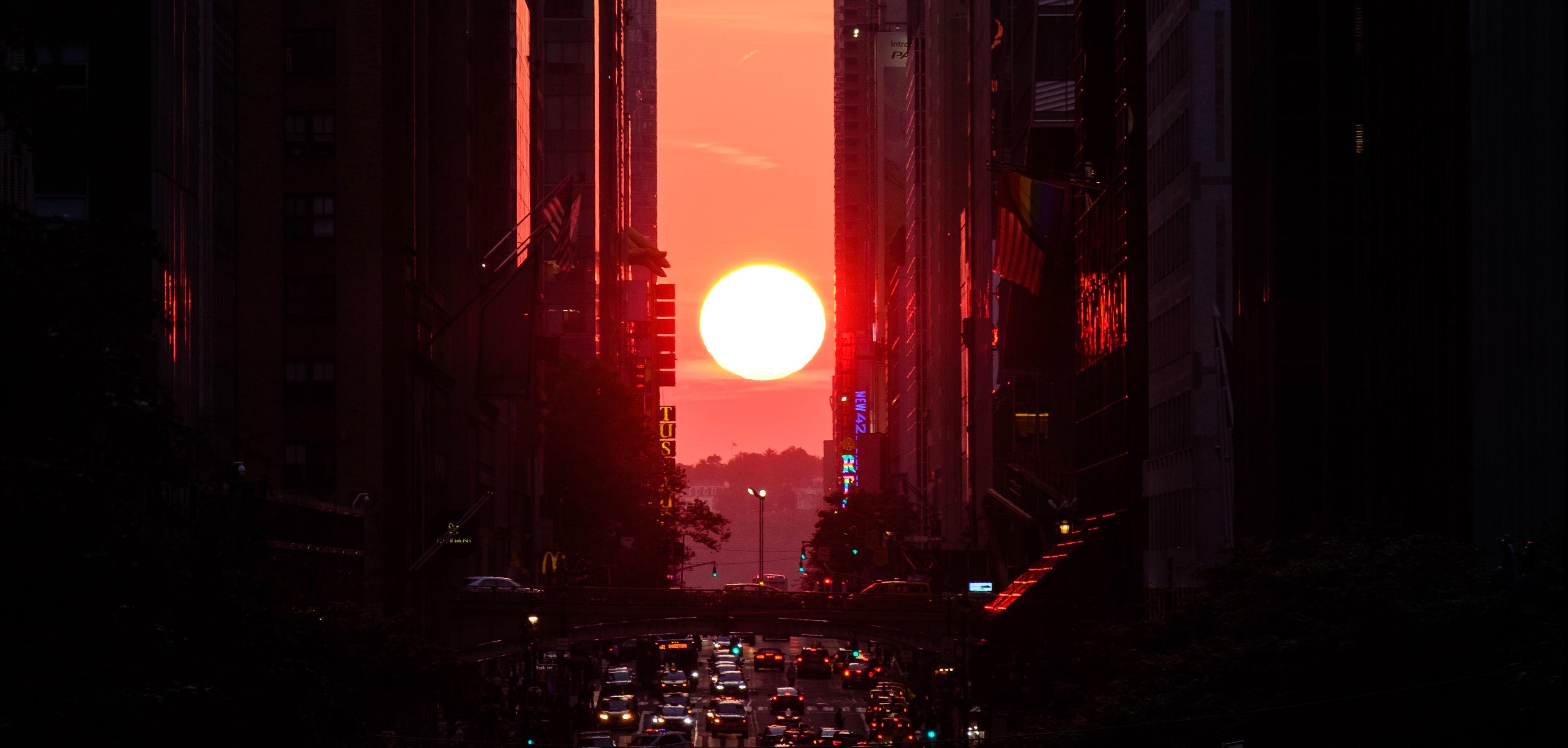 A sunset between buildings and a road full of vehicles.
