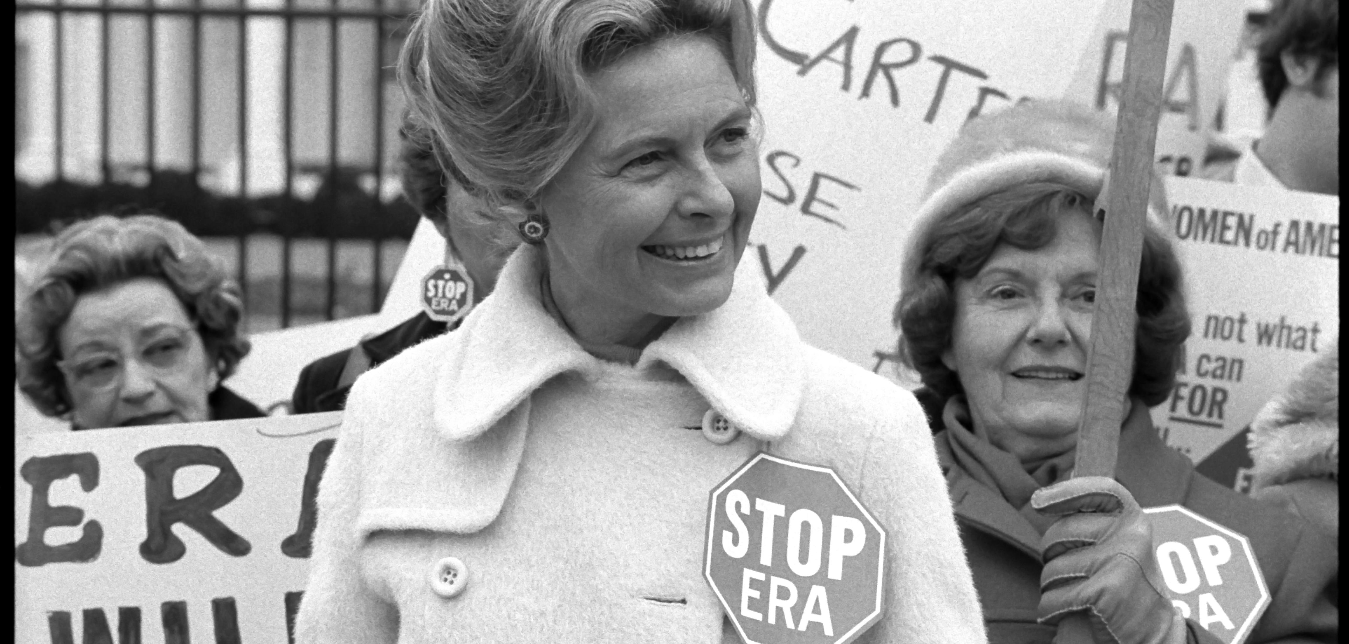 A White woman in a heavy coat wearing a "Stop ERA" badge smiles with other protesters in front of the White House fence. 