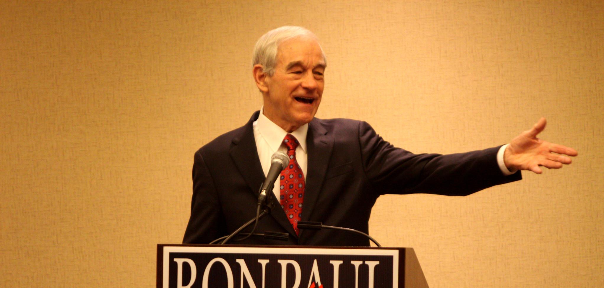 A man in a suit talking into a mic and pointing to his left. A sign on the podium says "Ron Paul, 2012, Restore America Now"