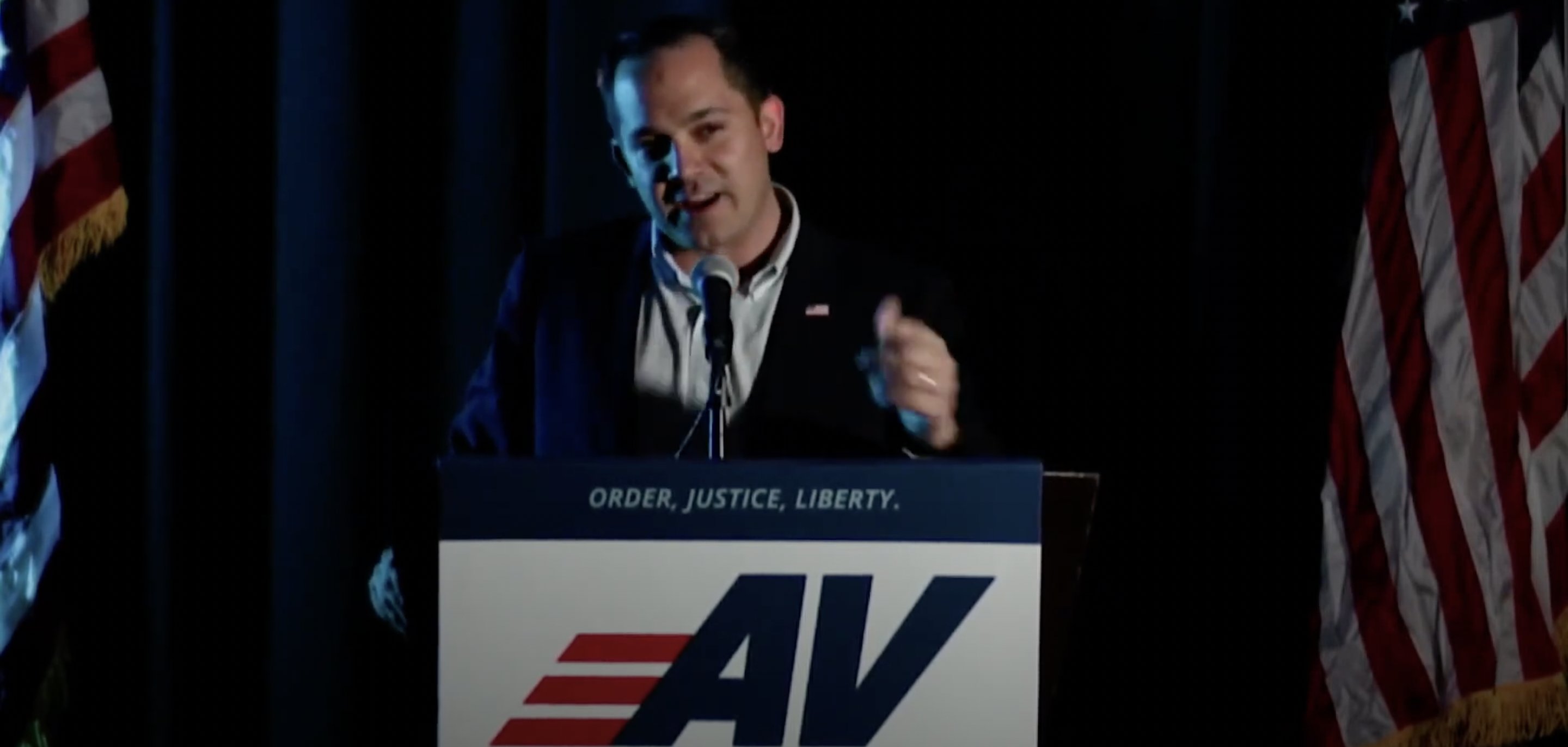 A man in blue suit standing at a podium with the letters AV on it speaking into a mic with two American flags behind him.