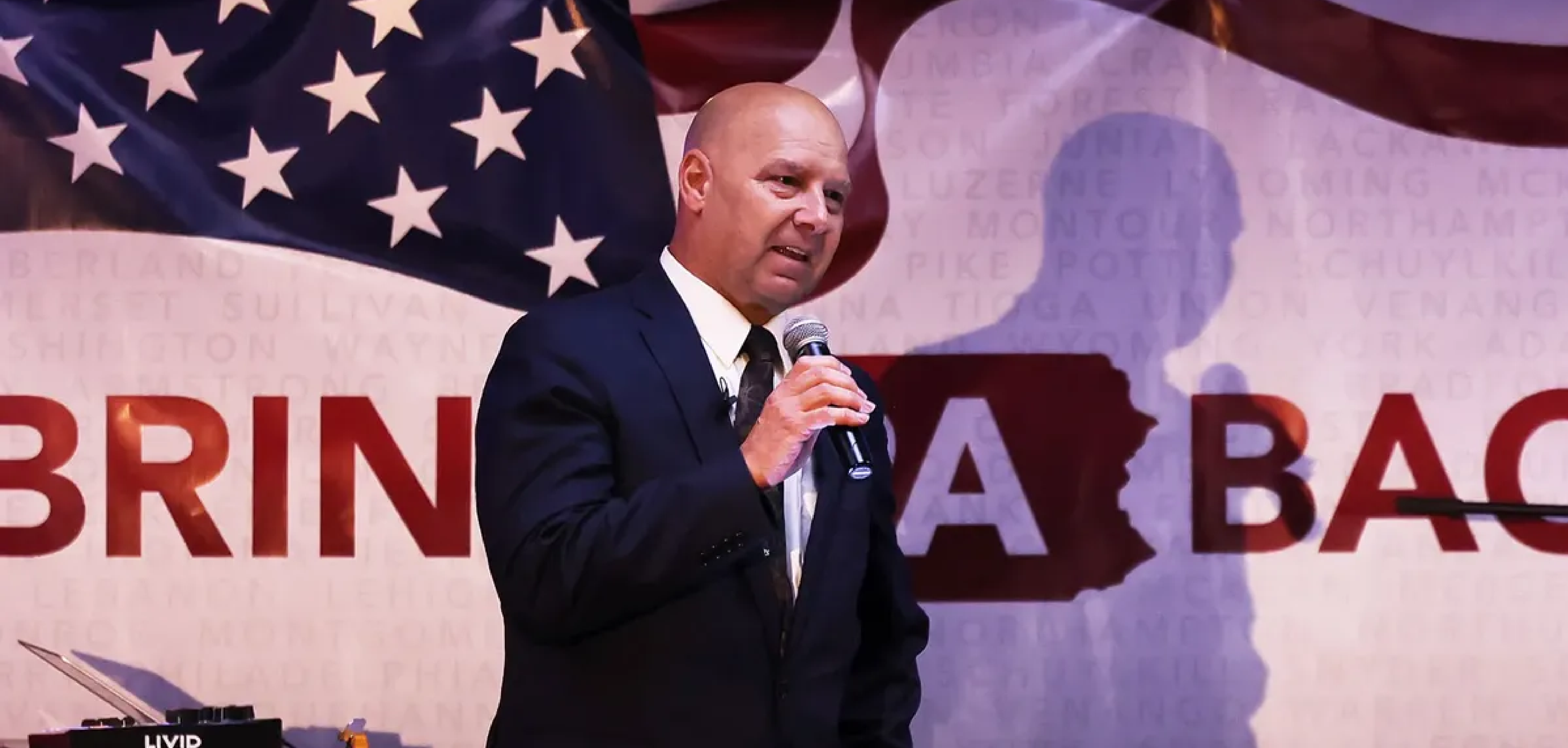 A bald man in a suit, standing in front of an American flag, talking into a mic.