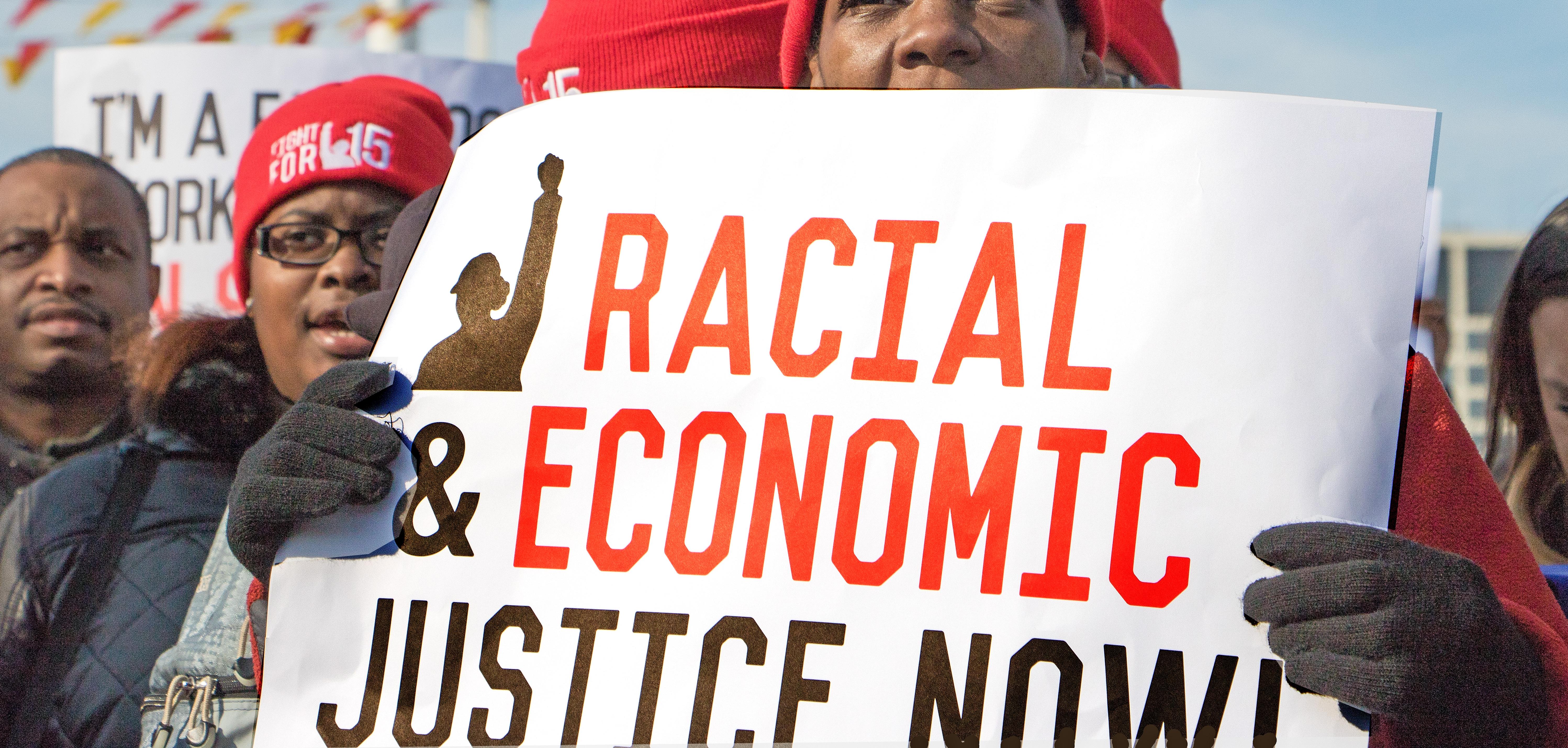 Woman in hat and gloves holds sign reading "Racial Economic Justice Now."