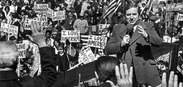 Jerry Falwell, a white man wearing a suit coat, speaks in front of a crowd holding signs reading "I love America."