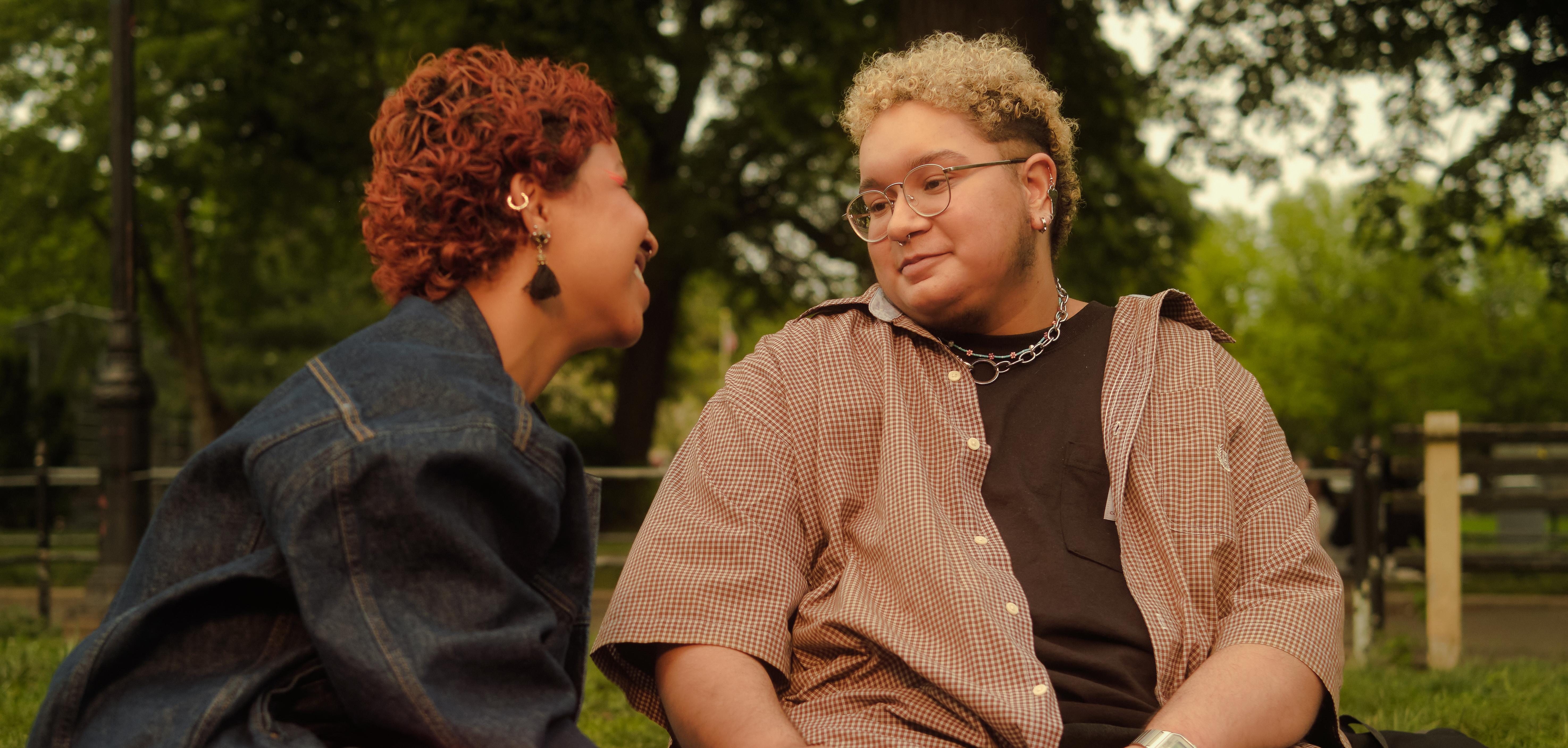 Two trans people in a park, looking at each other, laughing.