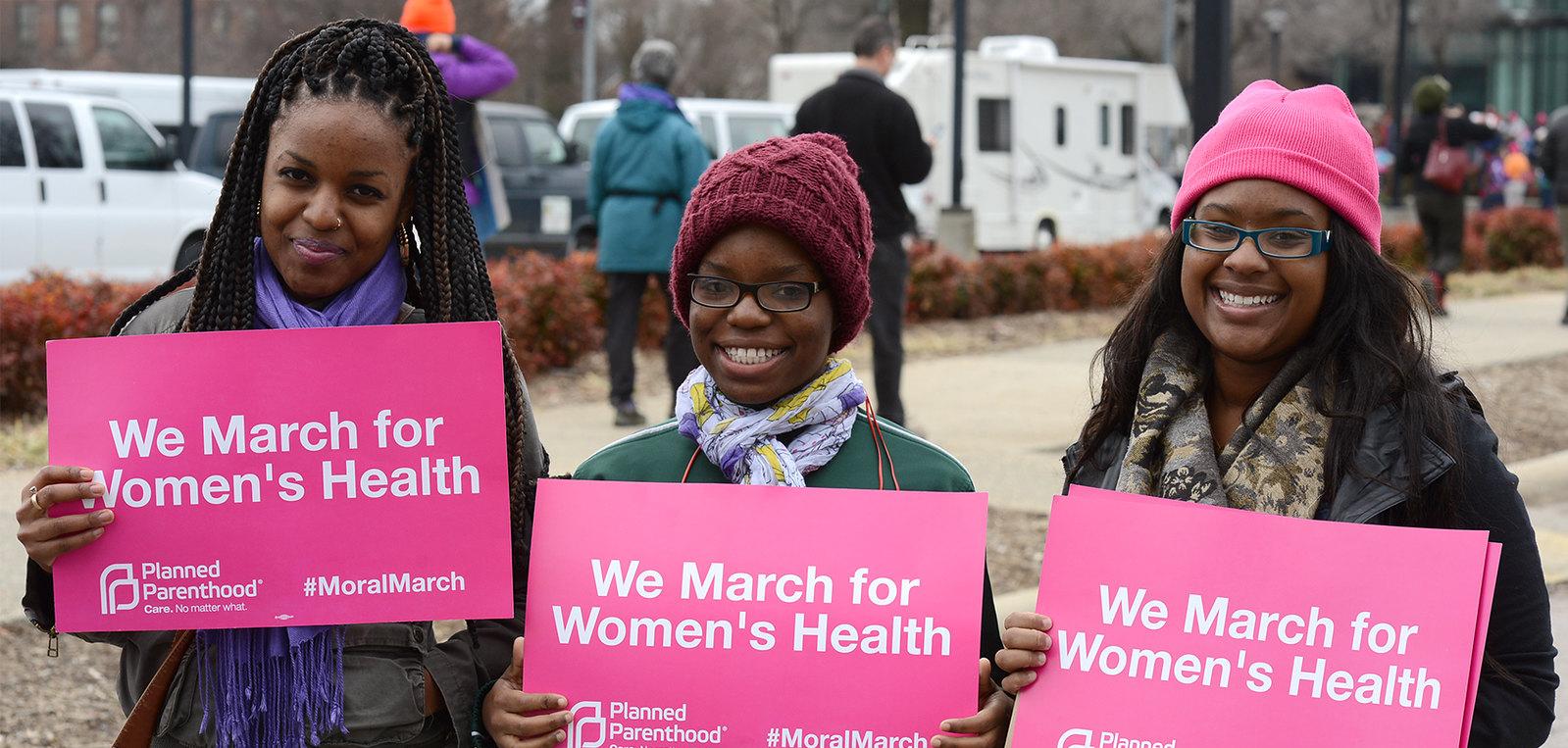 Three Black women wearing winter clothes holding signs that say "we march for Women's Health"