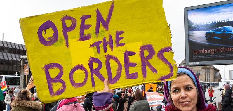 A woman with a scarf holding a sign that says "open the borders"