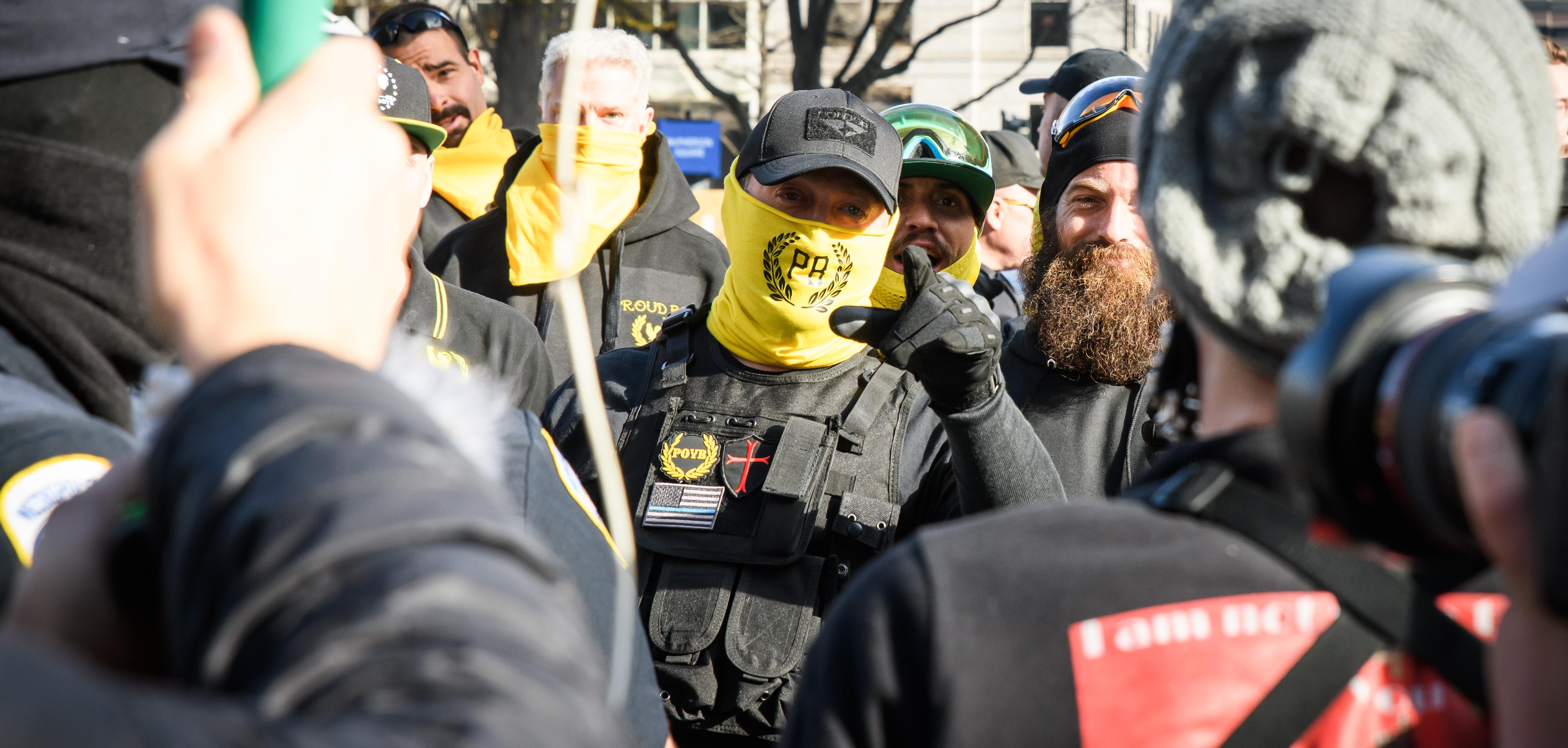 A Proud Boy with a neck warmer covering his mouth points mid-speech at the camera