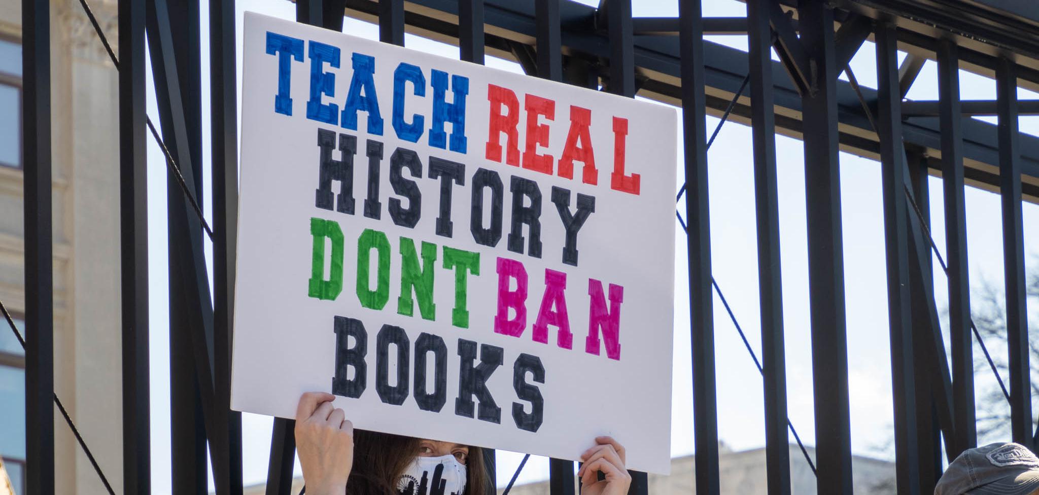Woman in blue shirt holding a sign that reads "Teach real history don't ban books"