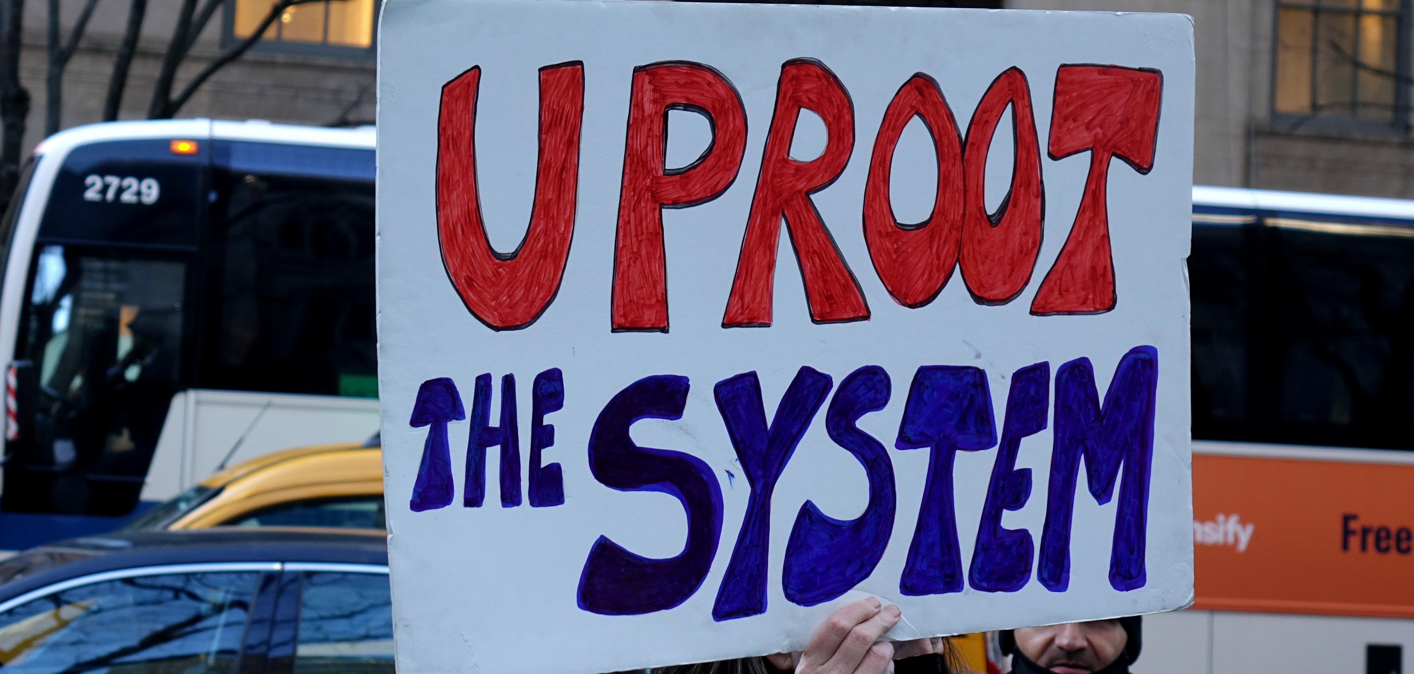 A person holds up a sign that reads "Uproot the system"