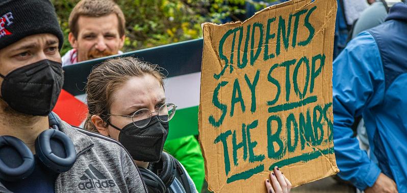 A person wearing a mask and glasses holds a sign reading "Students say stop the bombs"