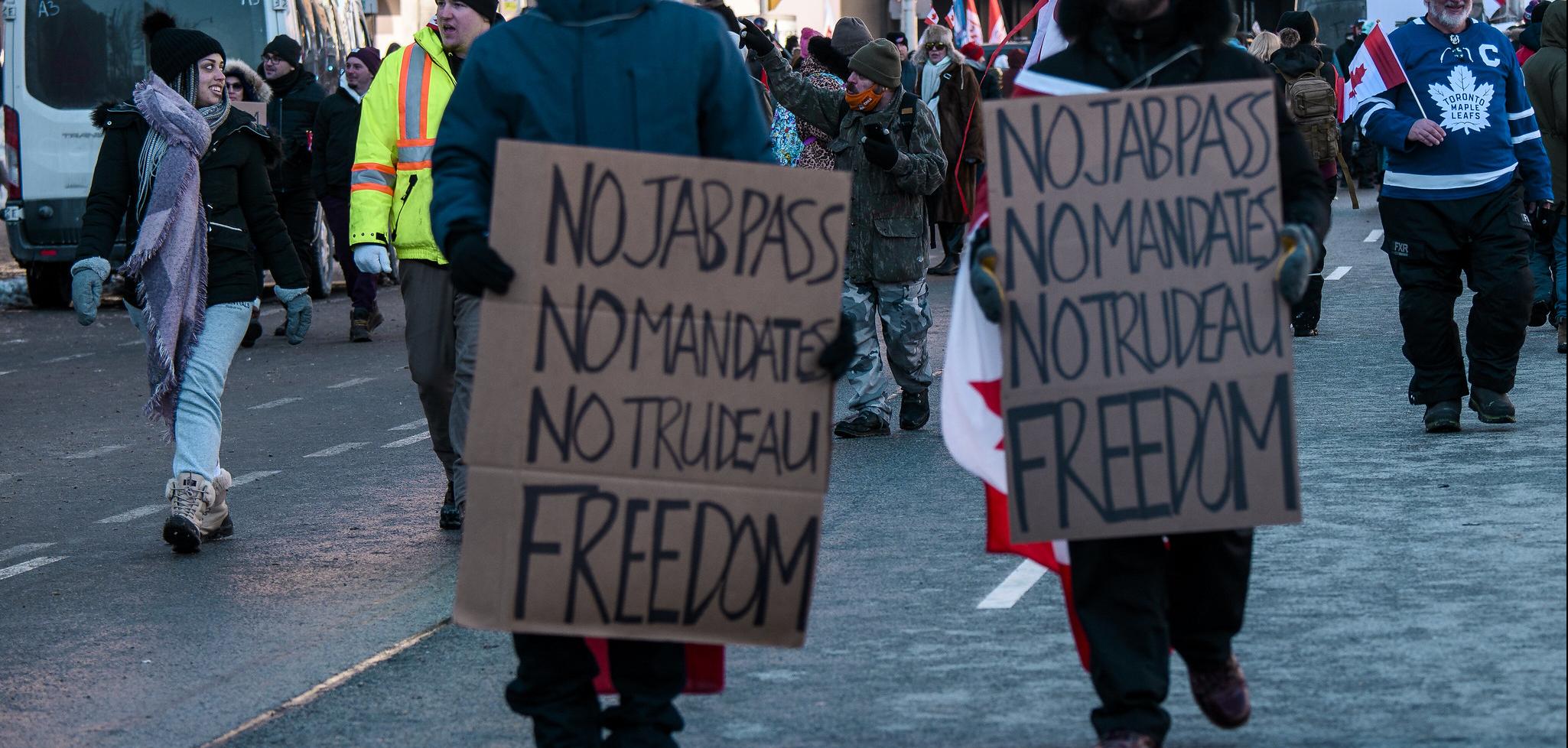 Two people in winter clothes holding signs that say "No jab pass, no mandates, no Trudeau, Freedom"