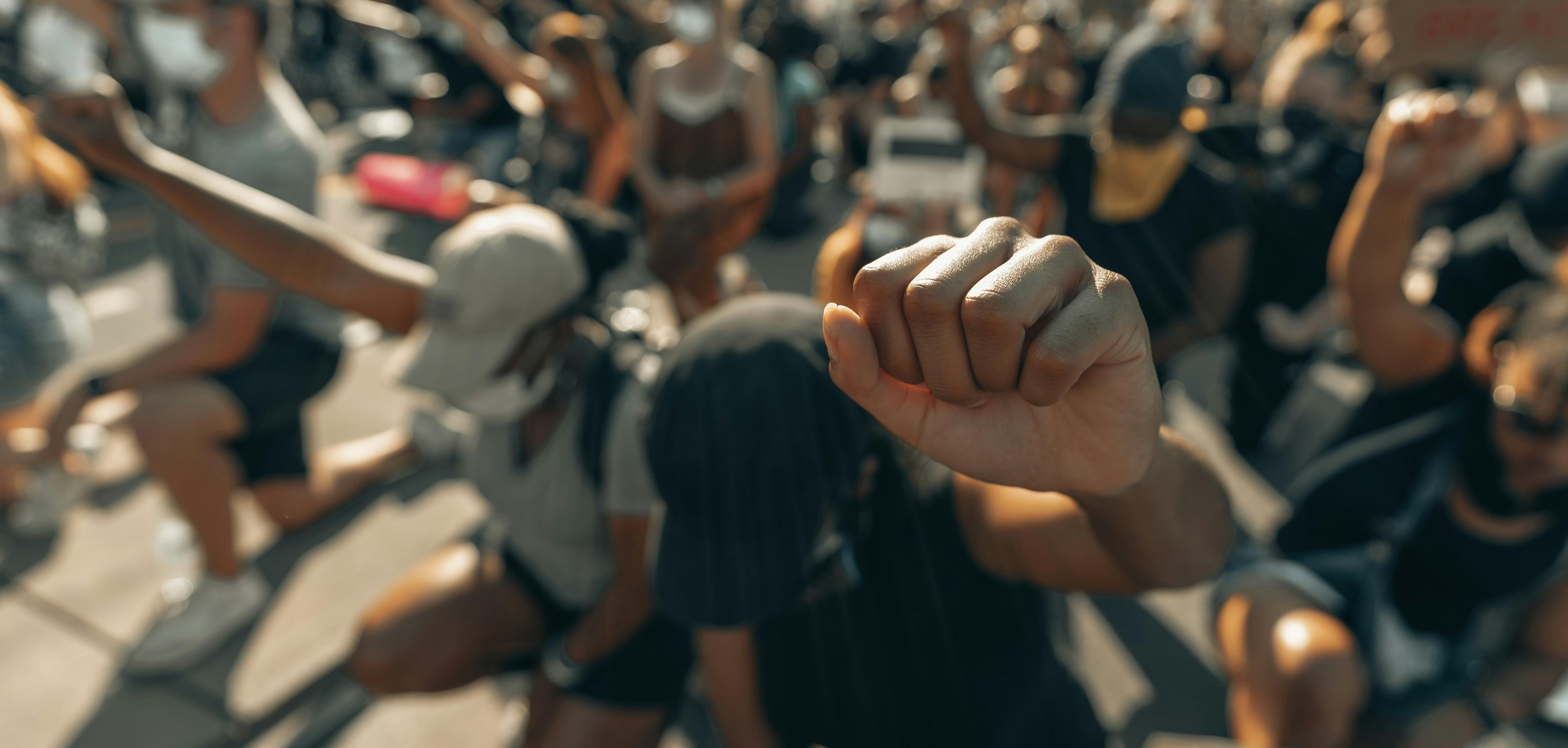 Focus on a raised fist, background of people kneeling in protest.