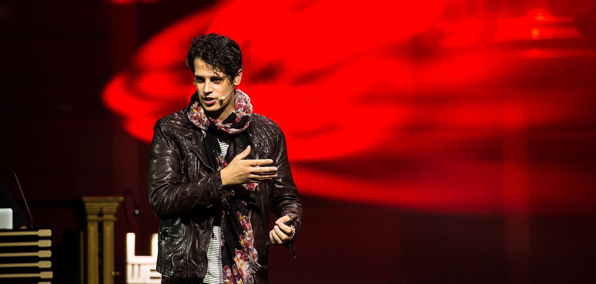 A man in a black jacket speaks in front of a red blurred background 