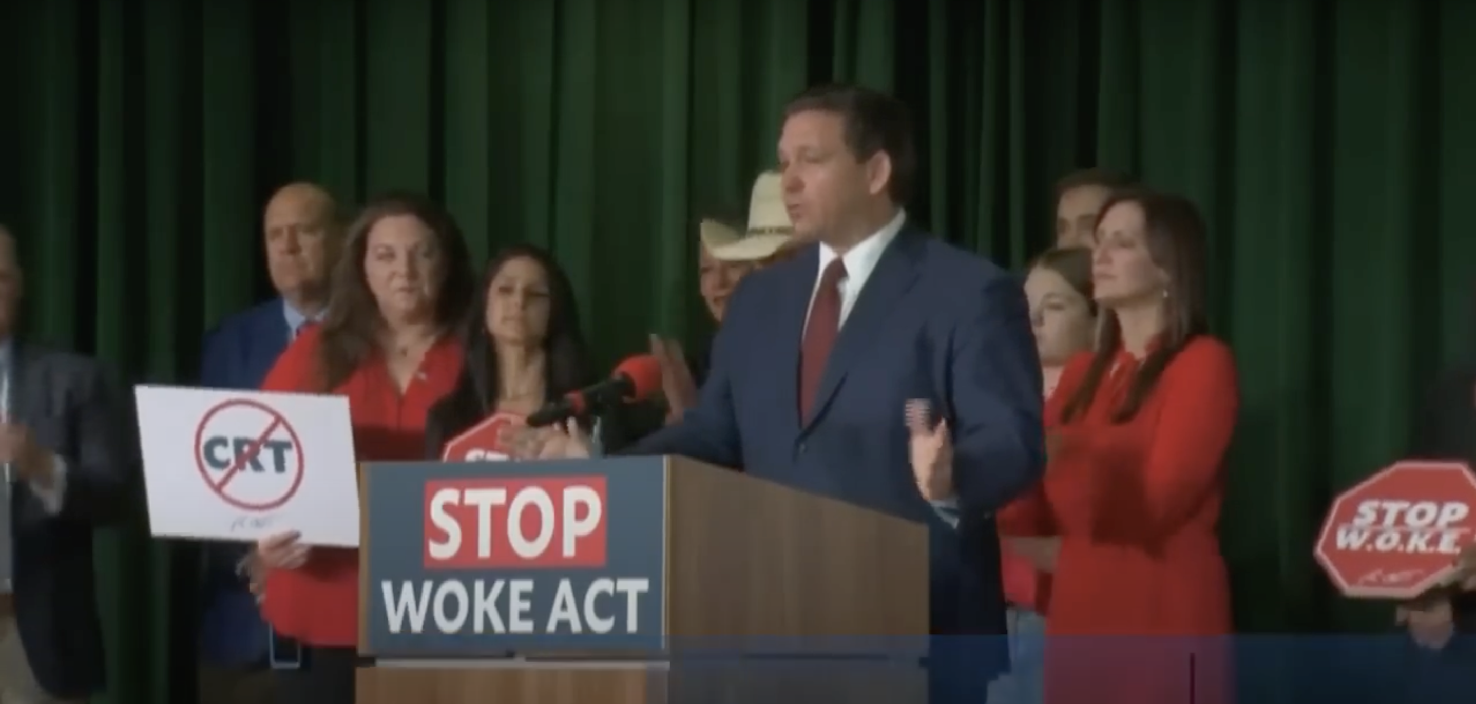 a man in a blue suit standing behind a podium with a sign that says "stop woke act"