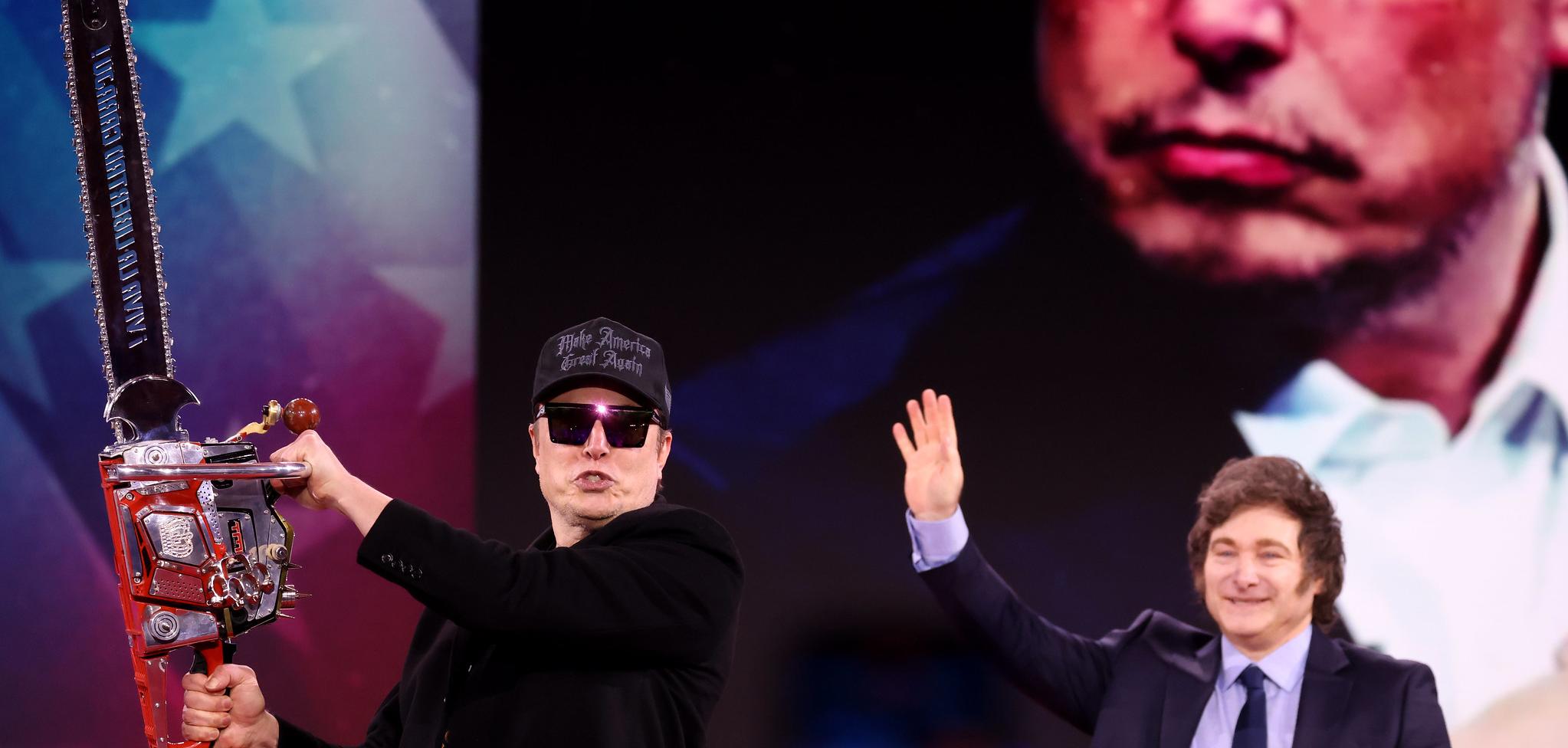 Two men standing in front of projector image. One man is waving at the crowd, the other man is holding a chainsaw