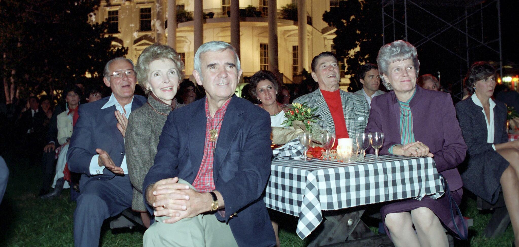Six people sitting around a gingham table cloth on a picnic table with the white house in the background
