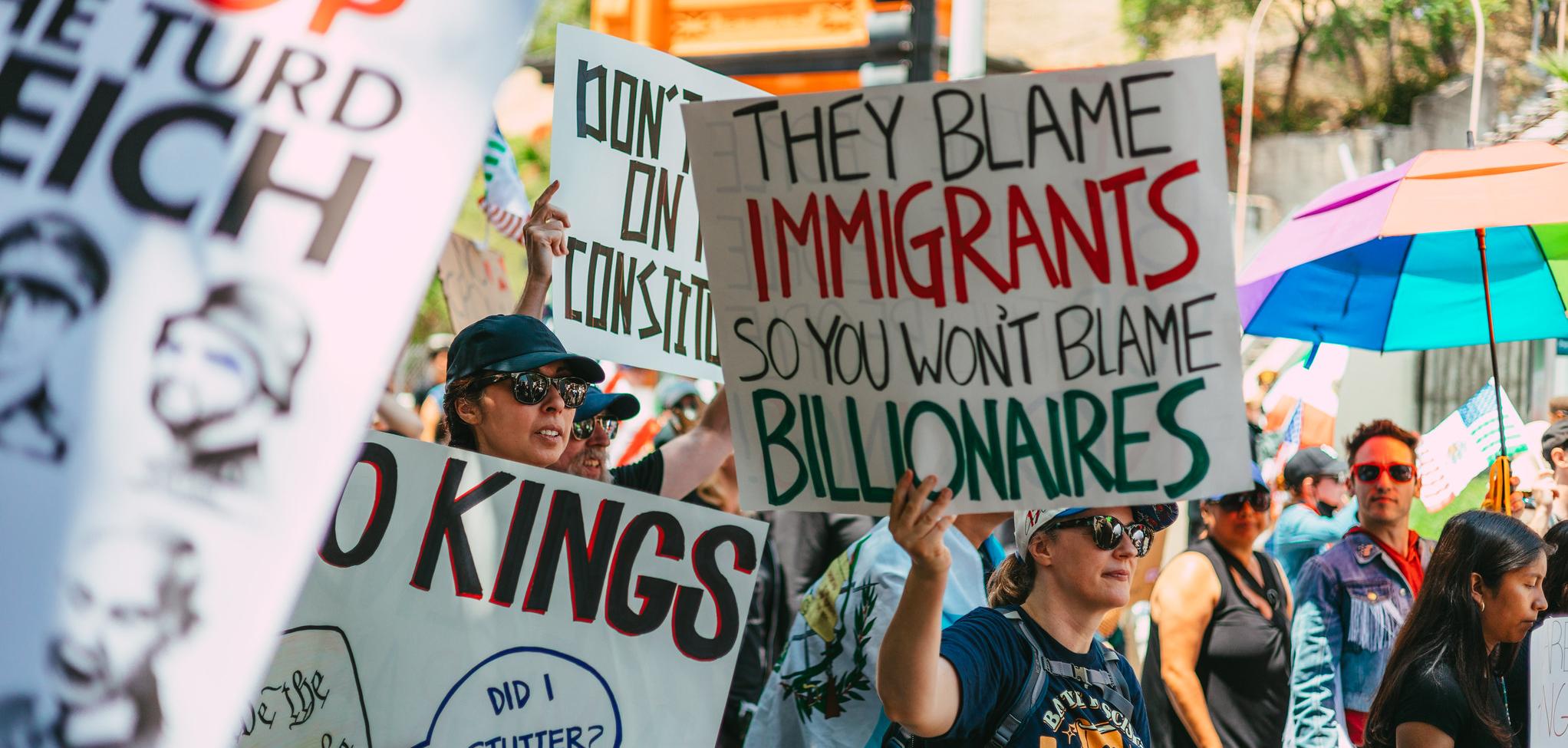 a person holding a sign that says "they blame the immigrants so you won't blame billionaires"