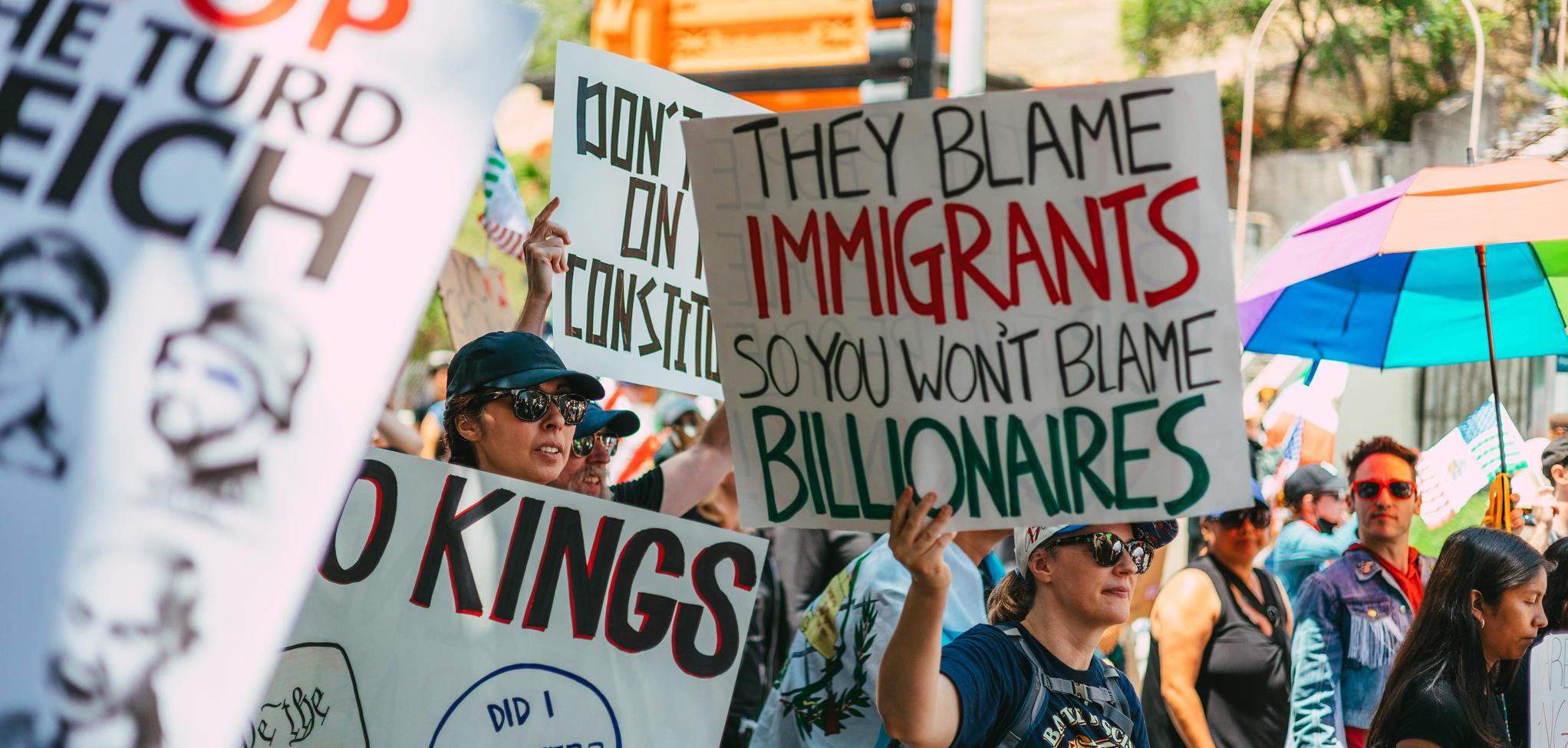 A protestor holding a sign saying "The blame immigrants so you won't blame billionaires"