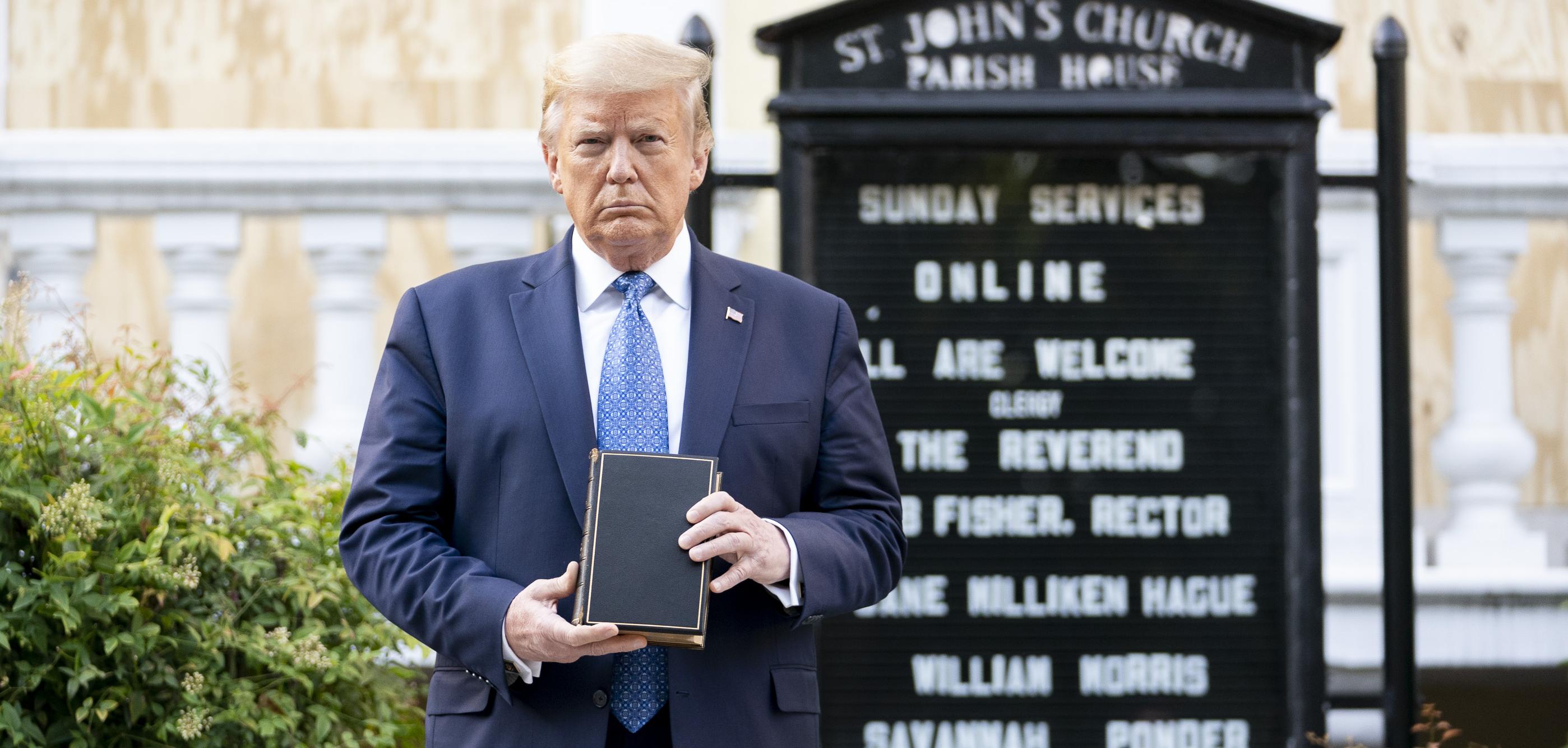 donald trump stands in front of a black sign for a church holding a bible in front of him
