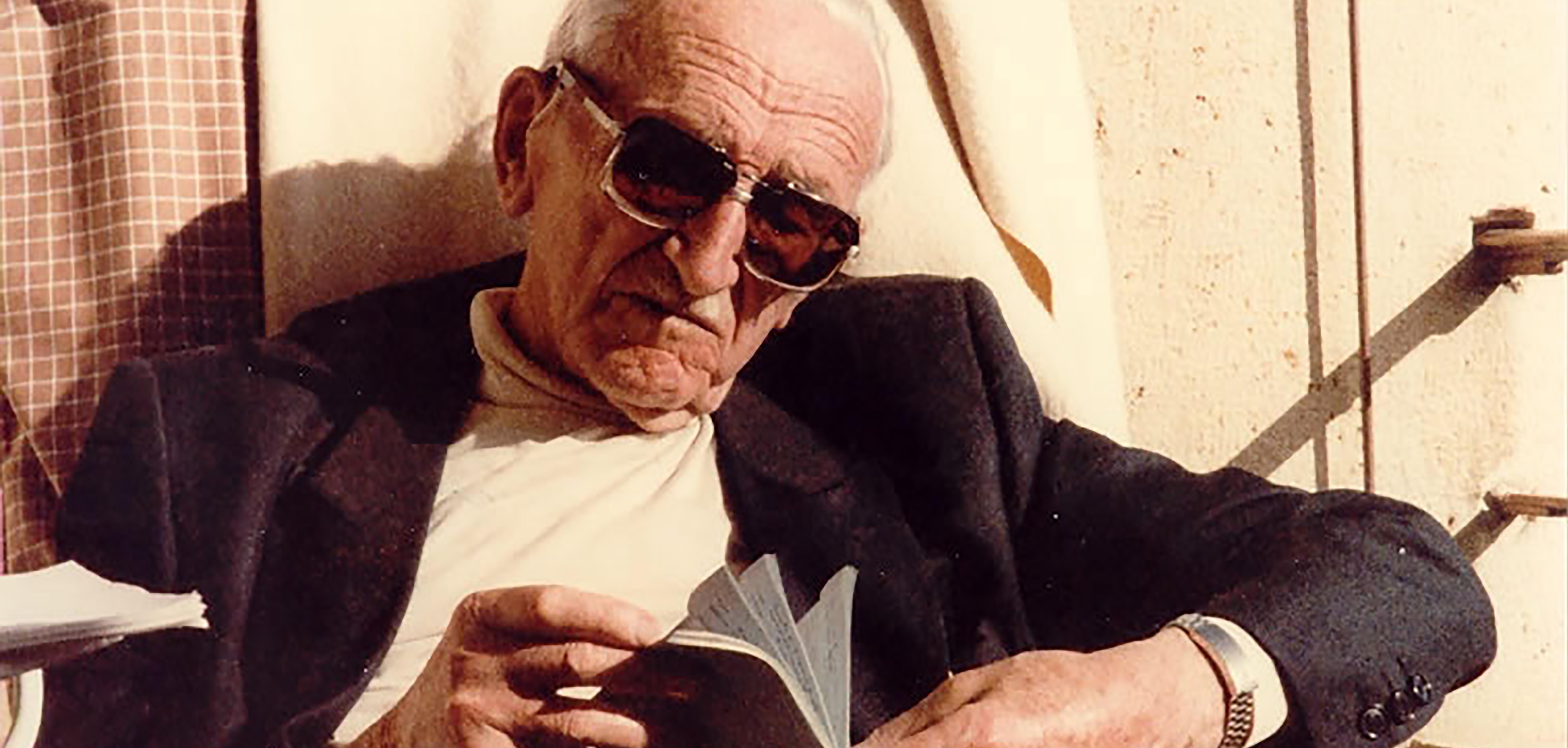 old man with sunglasses and white hair reads a book in the sun