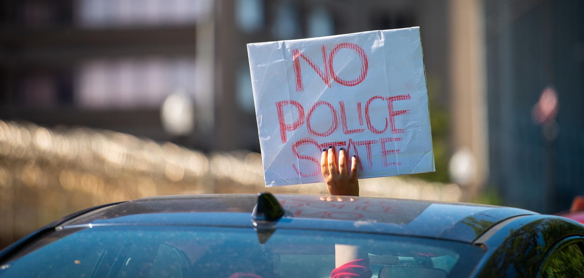 a person holding a sign out of the sunroof of their car that reads "no police state."
