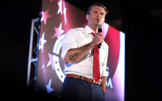 Pete Hegseth with a microphone standing in front of a projection of the American flag.