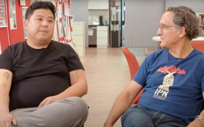Two men sitting in chairs, facing the camera but looking at each other.